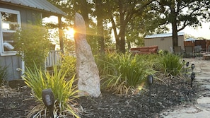 A garden area with a large rock, various plants, outdoor furniture, and a building with a metal roof.