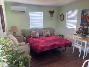 A compact room with a bed, a desk, and a chair. There is a window with blinds, a wall-mounted air conditioner, and a clock.