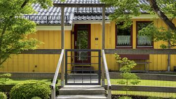 A building with a yellow exterior, a wooden staircase leading to a door, and a small garden area with a bench.