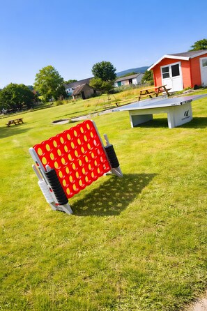 Un espace extérieur avec des sièges rouges et blancs, comprenant une table et des chaises, entouré d’un champ verdoyant et d’un bâtiment rouge.