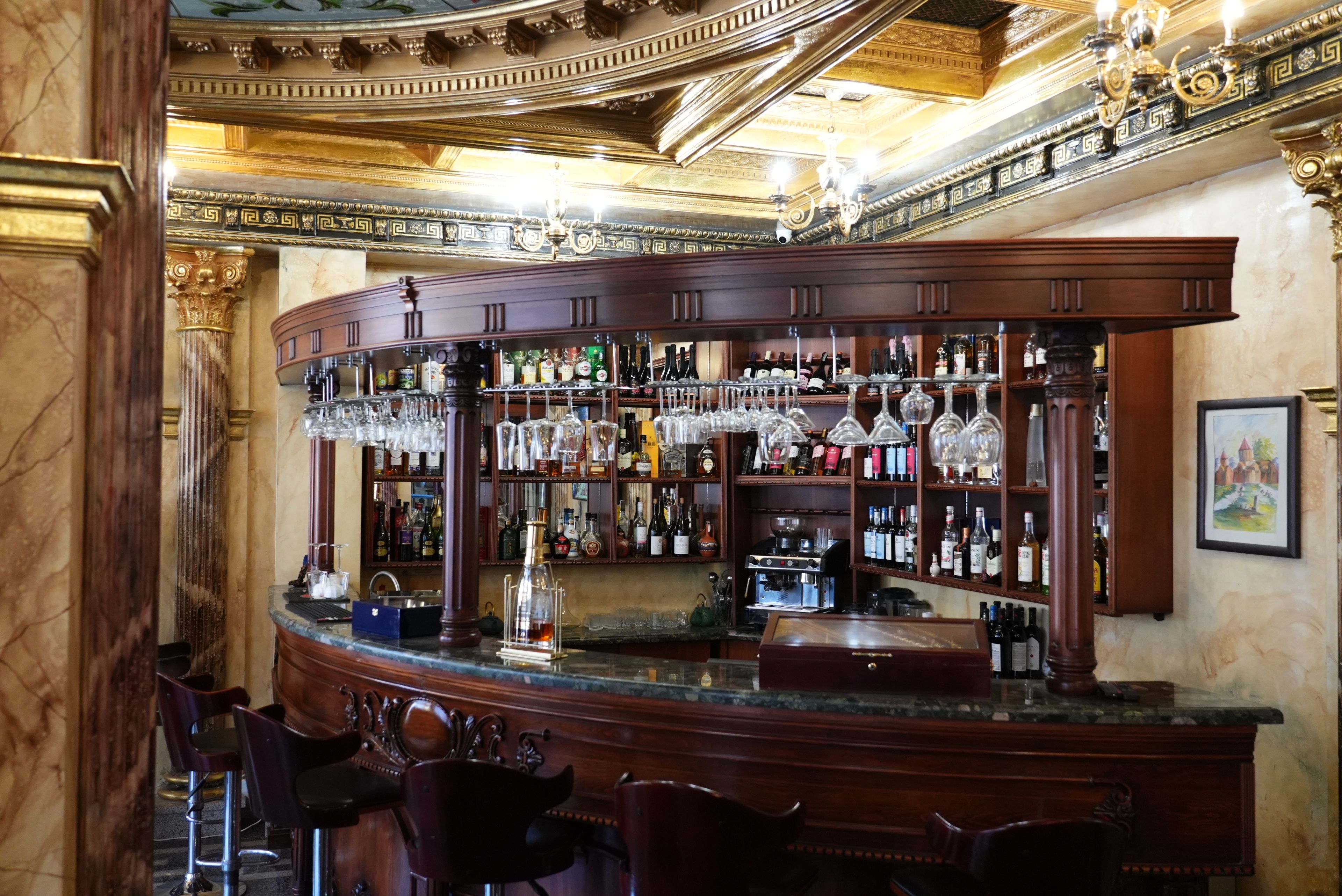 Uma área de bar com balcão curvo, prateleiras cheias de garrafas de bebidas e copos pendurados.