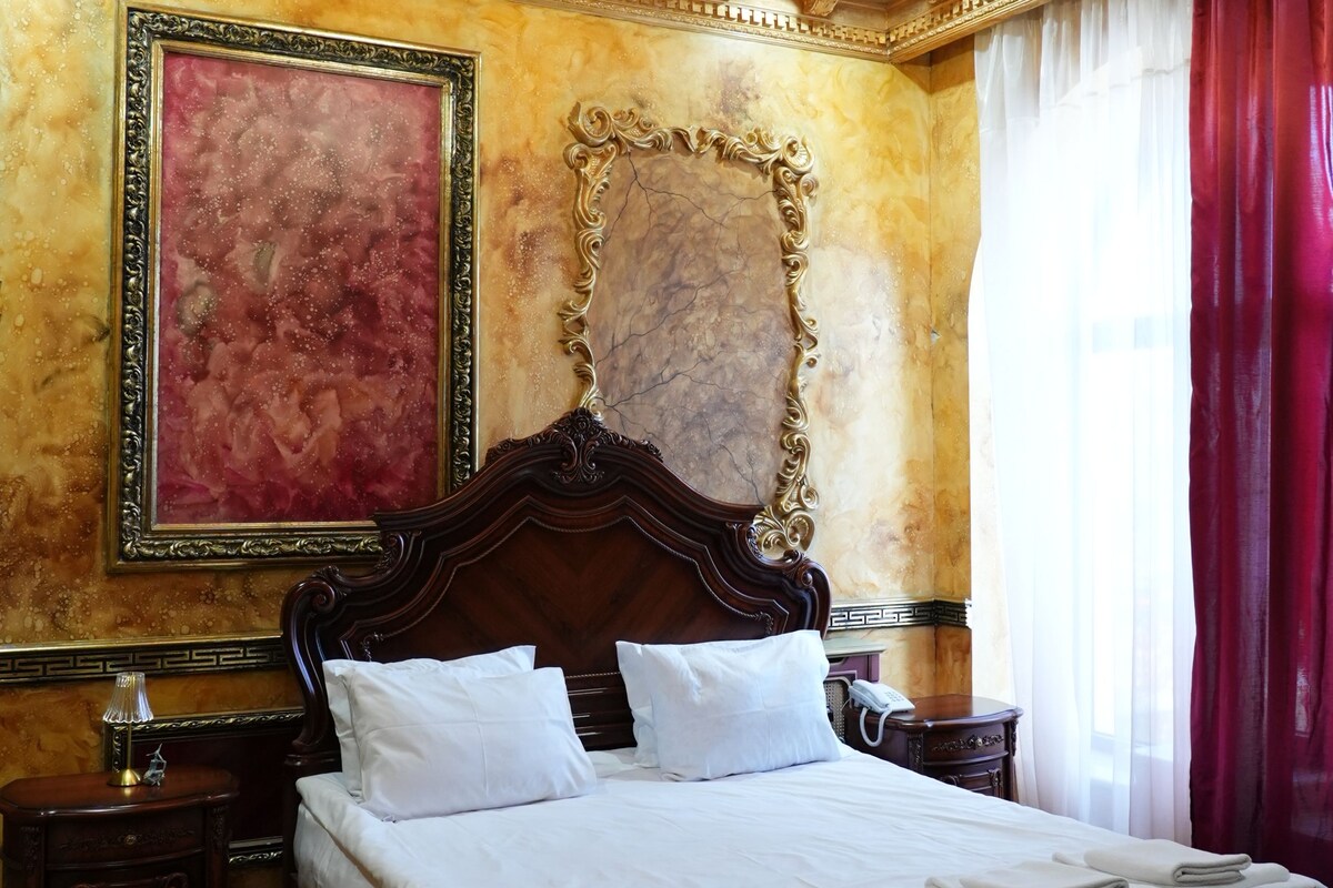 A luxurious bedroom with a large bed, ornate gold detailing, a chandelier, and a red curtain.