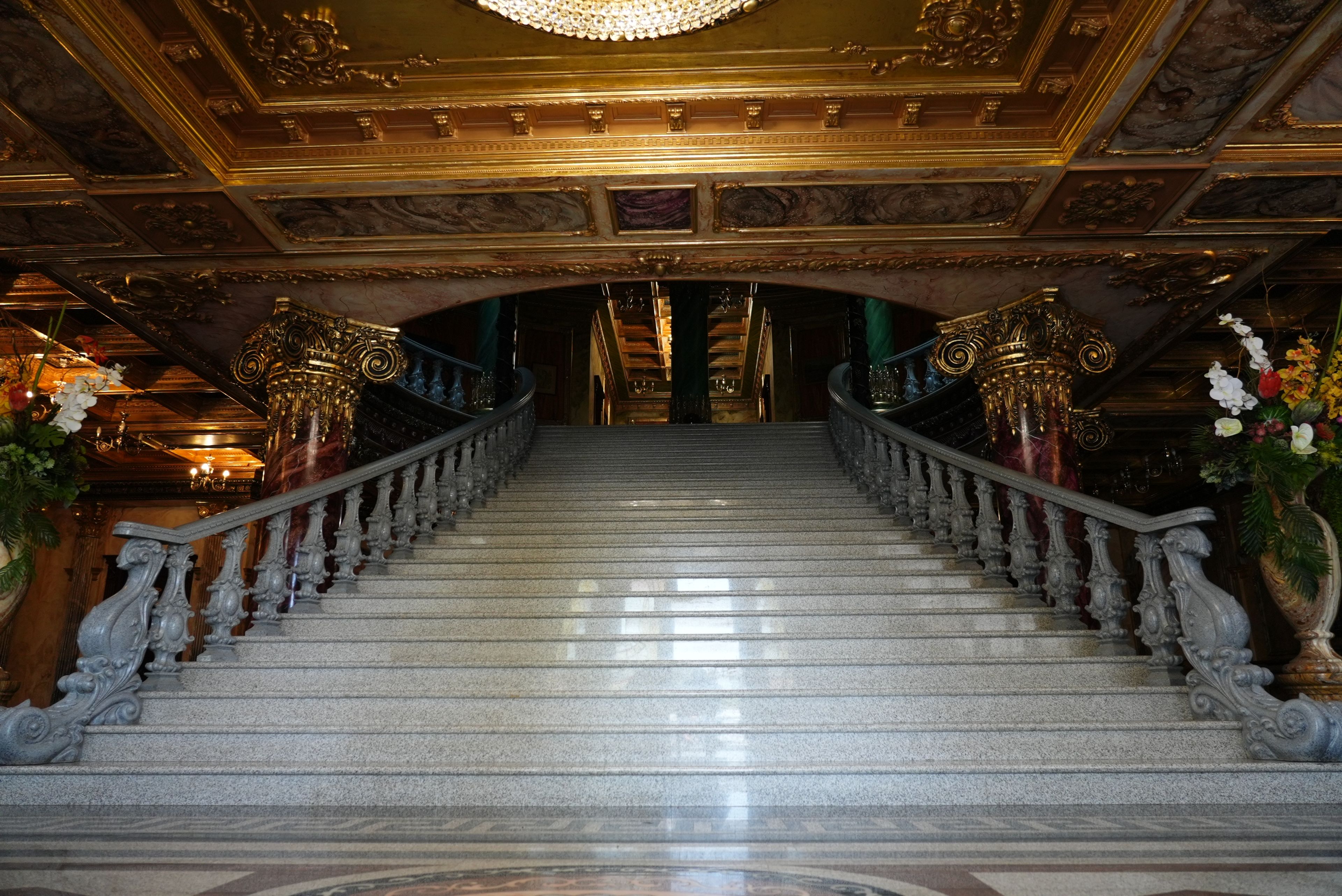 Una gran escalera con balaustradas ornamentales y un candelabro, que conduce a una habitación oscura con un techo alto.