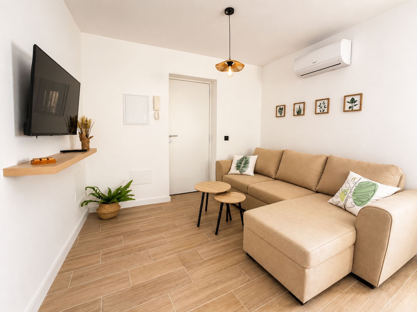 Een moderne woonkamer met een beige bank, een flatscreen televisie, een klein tafeltje en een potplant.
