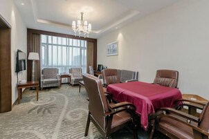 Quarto de hotel com mesa de jantar, cadeiras, lustre, televisão e uma janela grande.