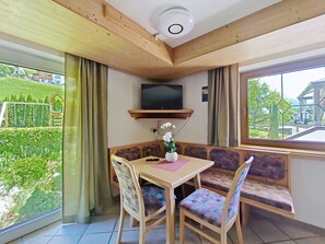 Ein Eckzimmer mit Holzbalkendecke, einem an einer Ablage montierten Fernseher, einem Tisch mit Stühlen, einer Bank und Blick auf eine grüne Landschaft durch große Fenster.
