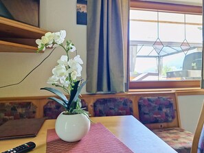 Una habitación con una mesa, un jarrón blanco con flores blancas, un control remoto y una ventana con vistas.