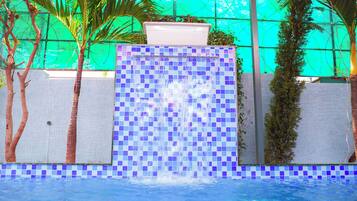 A swimming pool with a tiled waterfall feature, surrounded by palm trees and a green wall.