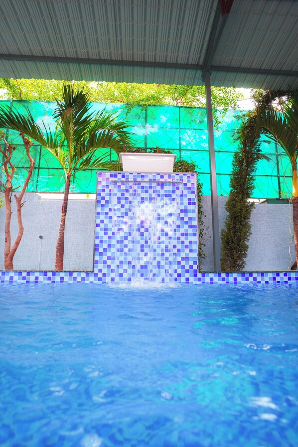 A swimming pool with a tiled waterfall feature, surrounded by palm trees and a green wall.