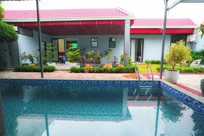 Una piscina con borde de azulejos, rodeada de plantas en macetas y un área de patio techada.