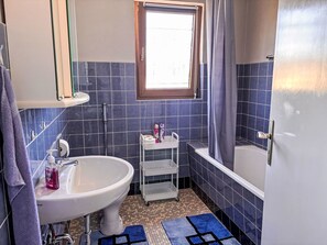 A bathroom with a white sink, a small white shelf, a window, a bathtub, and a shower curtain.