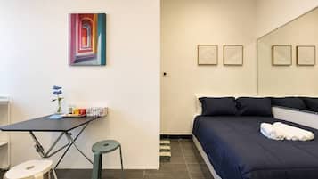 A hotel room with a bed, a desk with a chair, a small stool, and a painting on the wall.