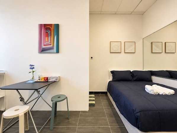 A hotel room with a bed, a desk with a chair, a small stool, and a painting on the wall.