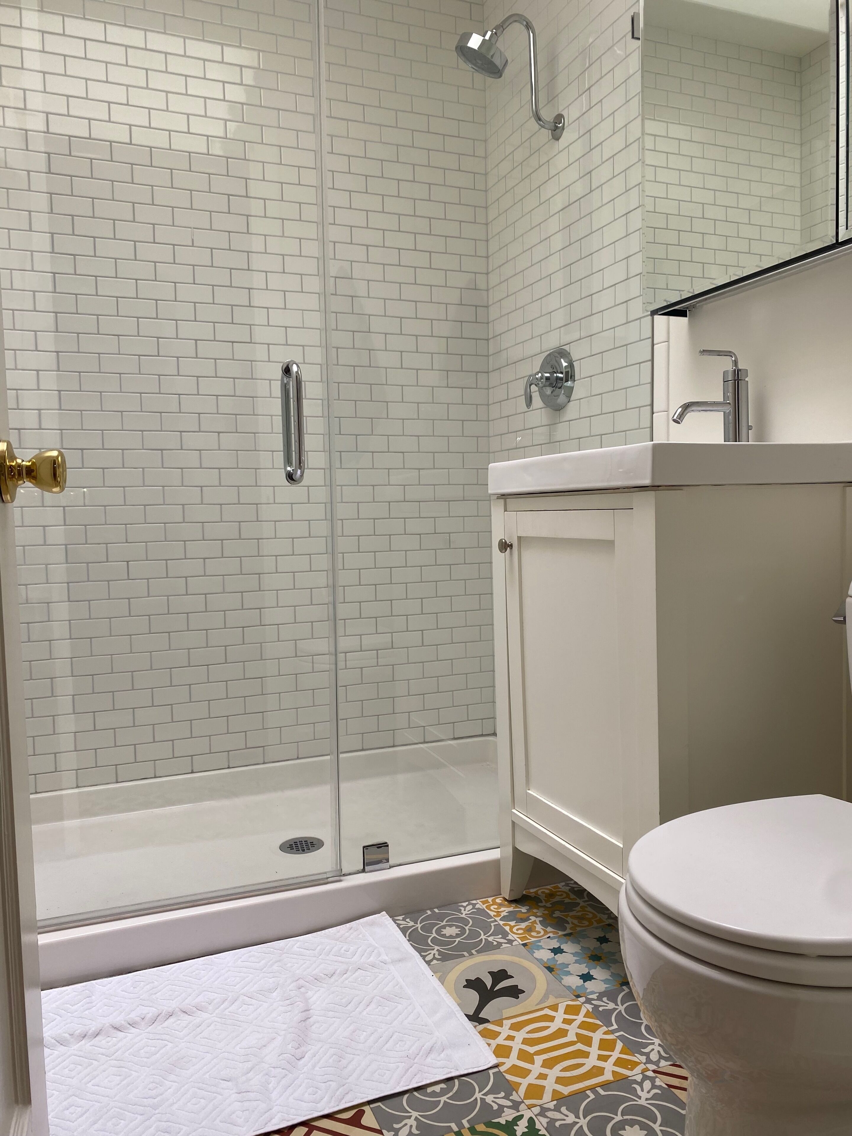 Un baño con paredes de azulejos tipo metro blancos, cabina de ducha de vidrio, inodoro blanco y piso estampado.