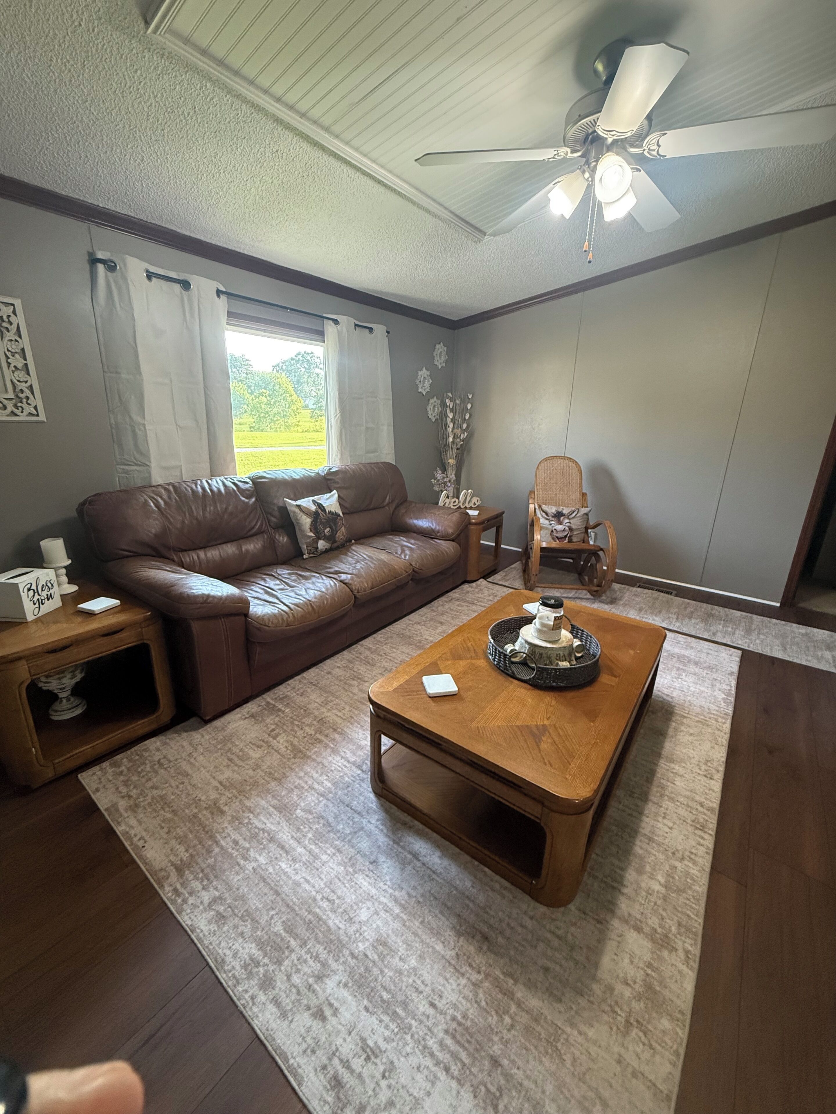 A living room with a brown leather sofa, a wooden coffee table, a ceiling fan, and a window with curtains.