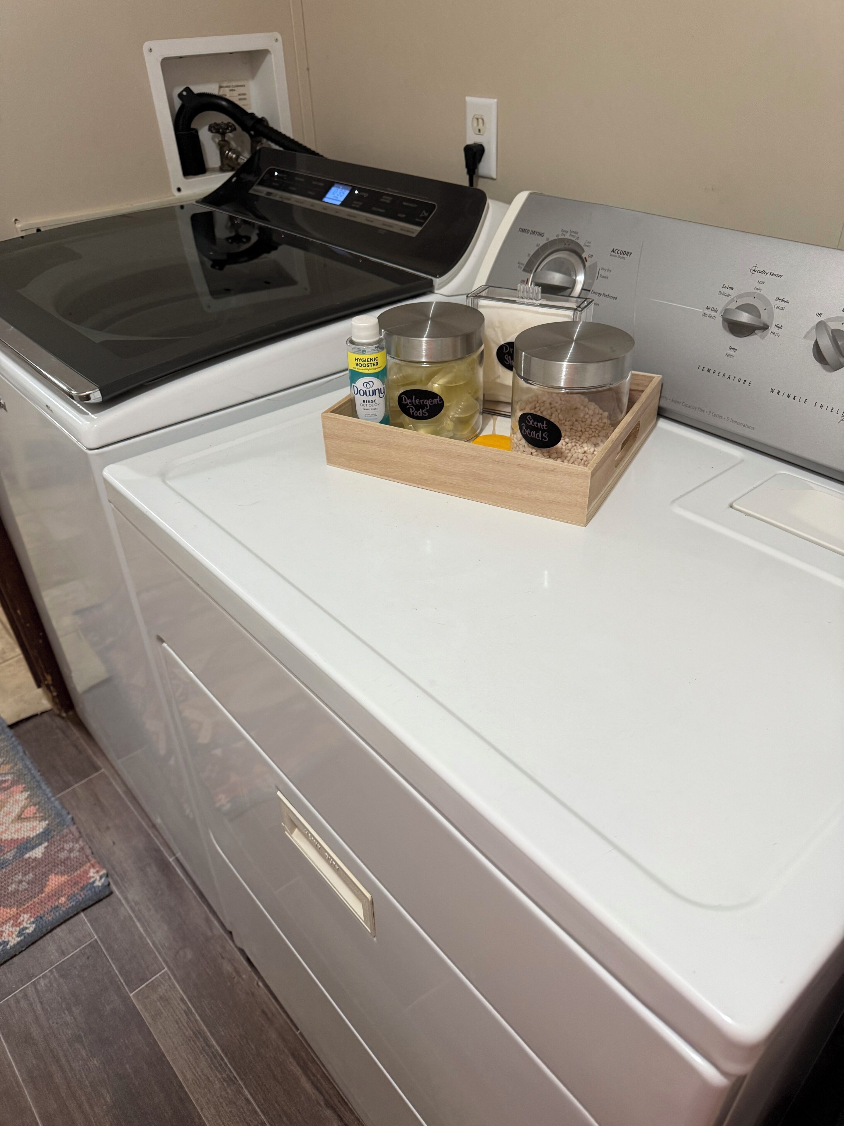 Una lavadora de carga frontal con un panel de control, una bandeja de madera con cápsulas de detergente, una botella de suavizante de telas y un recipiente con perlas perfumadas encima.
