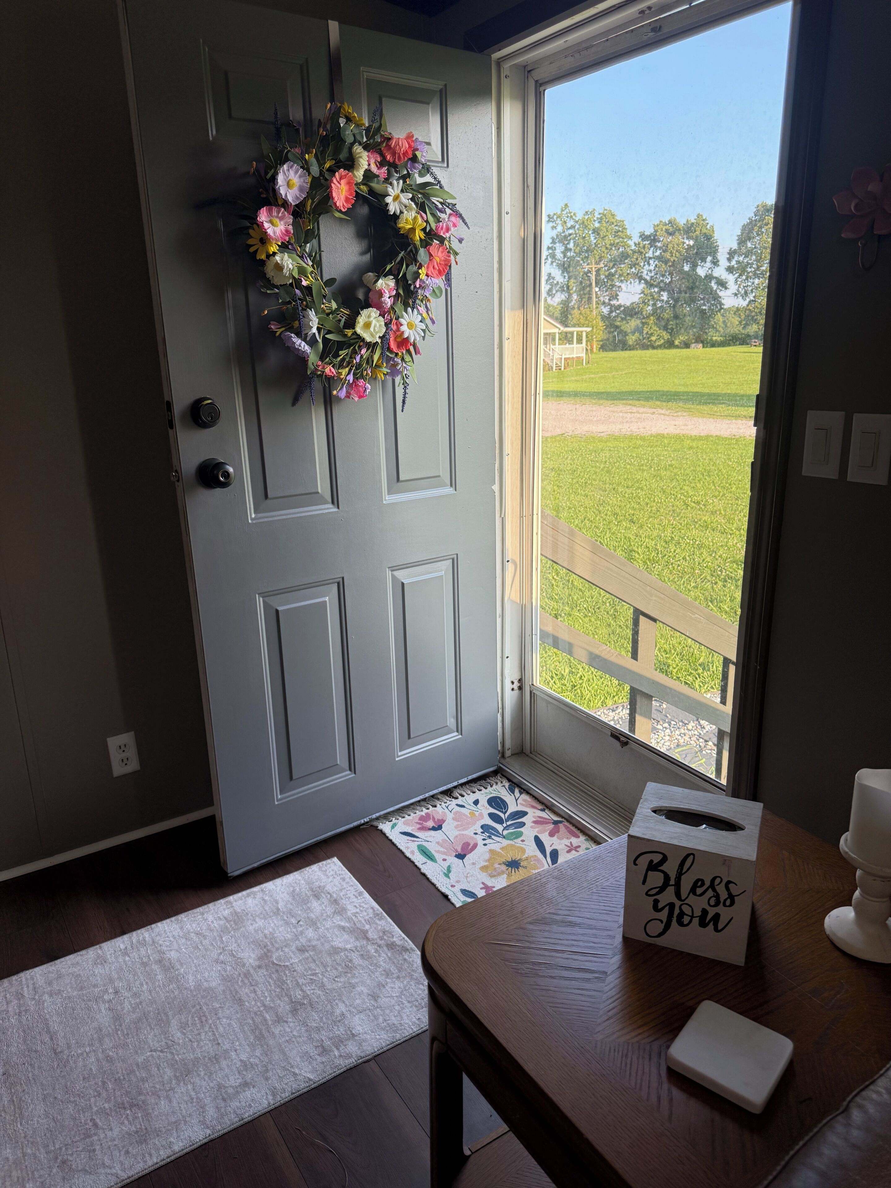 A door with a floral wreath, a wooden bench, a tissue box, and a view of a grassy field through a window.