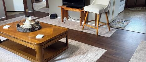 A living room with a wooden coffee table, a television on a stand, a white chair, and a kitchen area in the background.