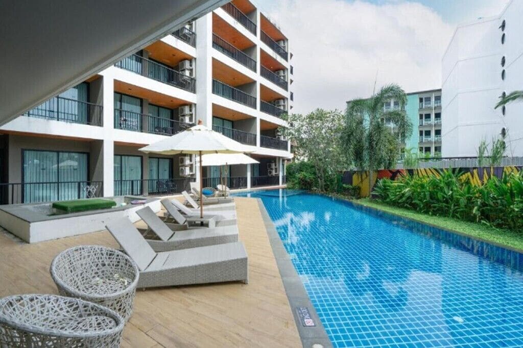 Piscina con sillas de descanso y una sombrilla, rodeada por un edificio de varios pisos con balcones.