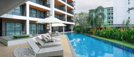 Piscina con sillas de descanso y una sombrilla, rodeada por un edificio de varios pisos con balcones.