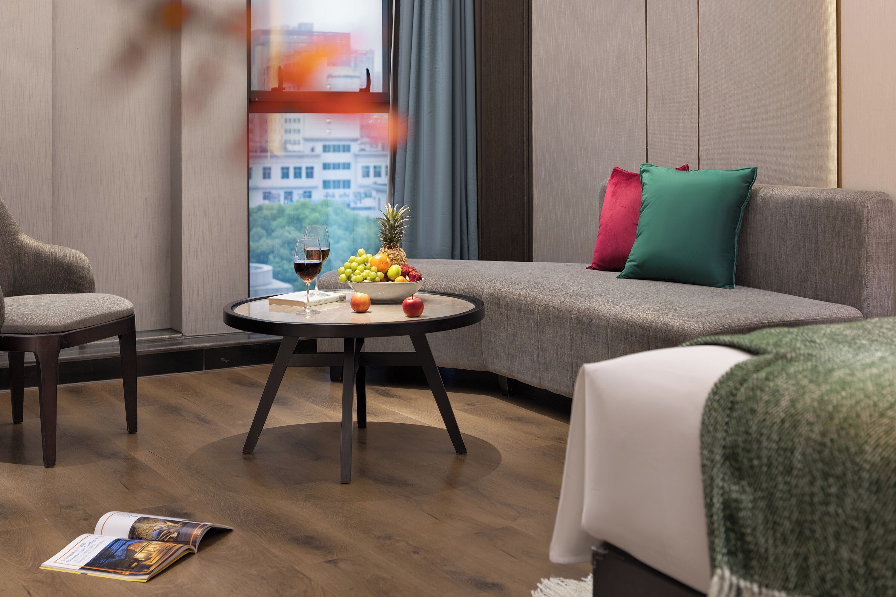 Une chambre d’hôtel moderne avec un canapé gris, une table ronde sur laquelle se trouve un panier de fruits et des verres à vin, et une vue sur le paysage urbain par la fenêtre.
