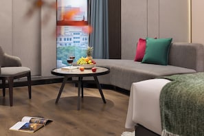 Una habitación de hotel moderna con un sofá gris, una mesa redonda con frutero y copas de vino, y vistas a un paisaje urbano a través de la ventana.