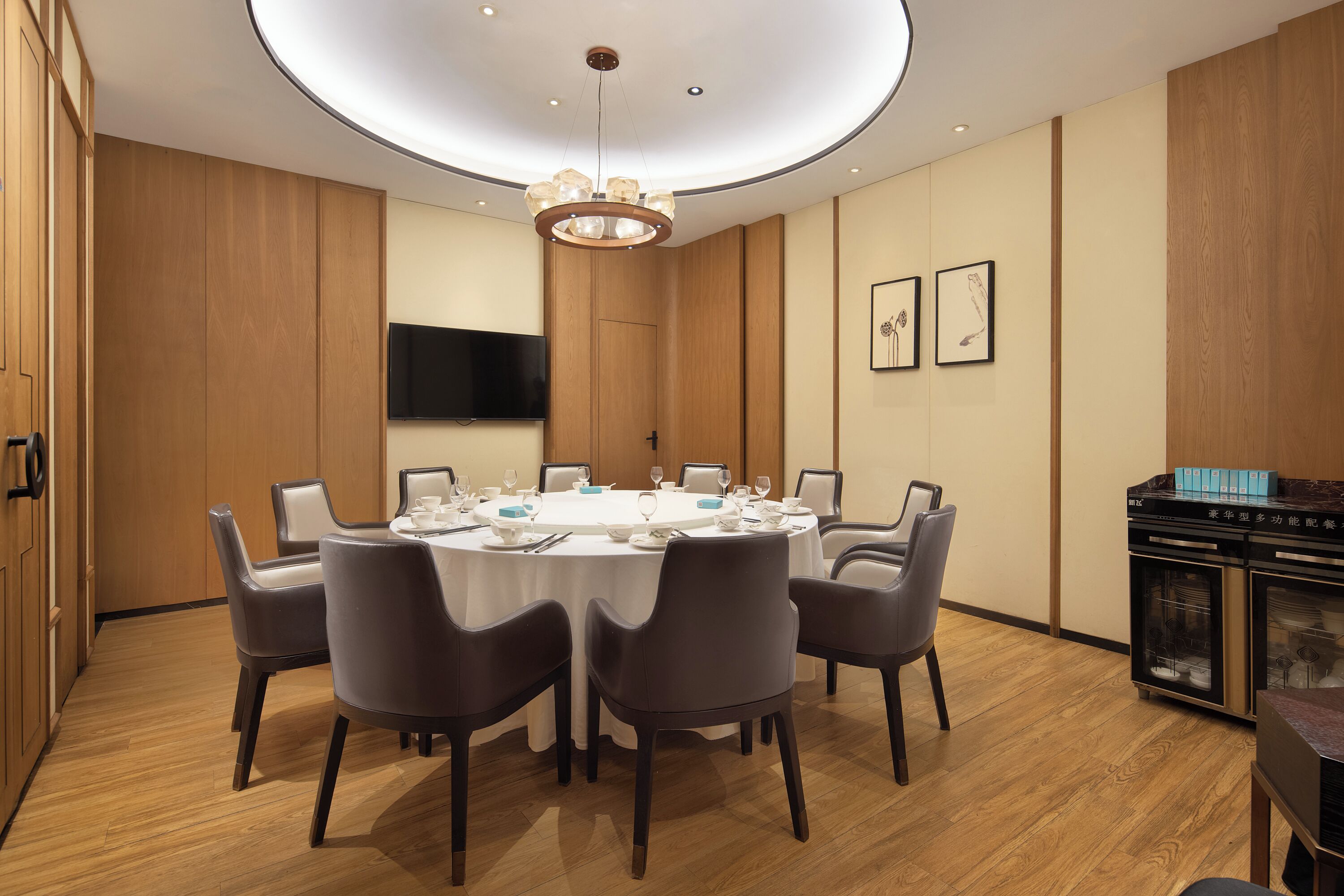 A dining room with a round table set for six, surrounded by chairs, a television mounted on the wall, and framed artwork above the table.