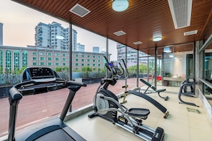 Un gimnasio con equipamiento de ejercicio, ventanas amplias y vista a edificios.