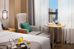 Habitación de hotel con una cama, una silla con un cojín, una mesita con tetera, un teléfono y vistas a edificios a través de la ventana.