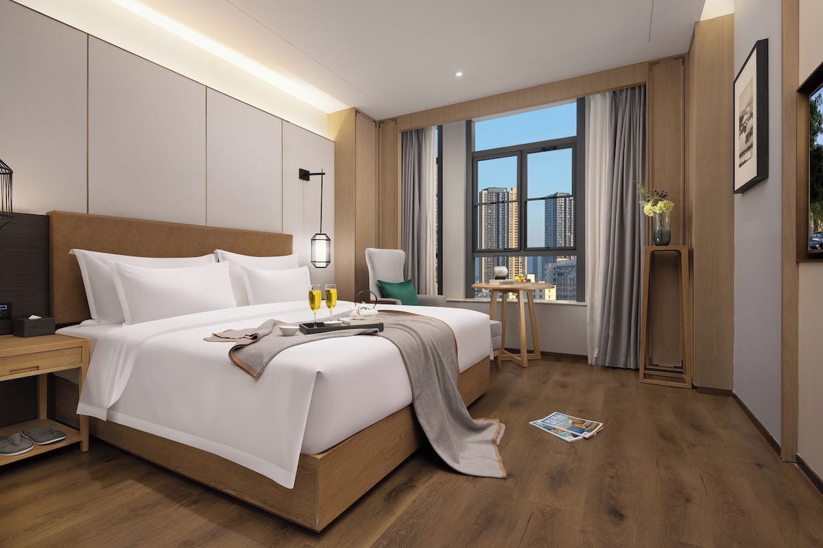Habitación de hotel con una cama grande, mesita de noche, una pequeña mesa con un florero y una ventana con vista a la ciudad.