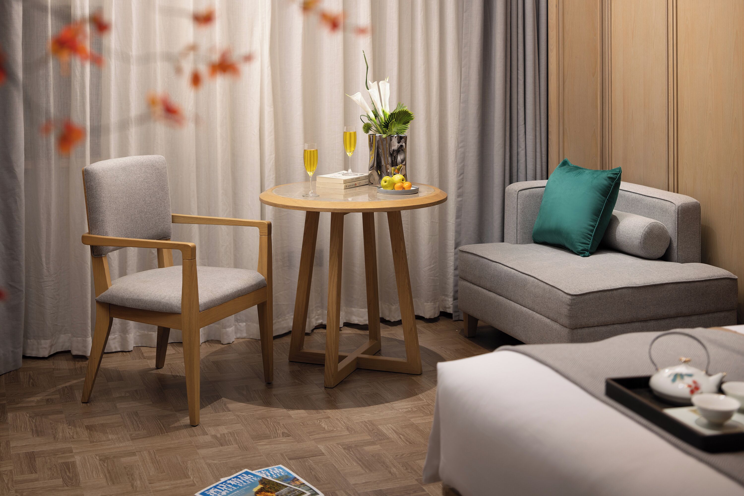 A hotel room with a wooden chair, a round table with a vase and books, a sofa with a teal pillow, and a bed with a tray of tea and cups.