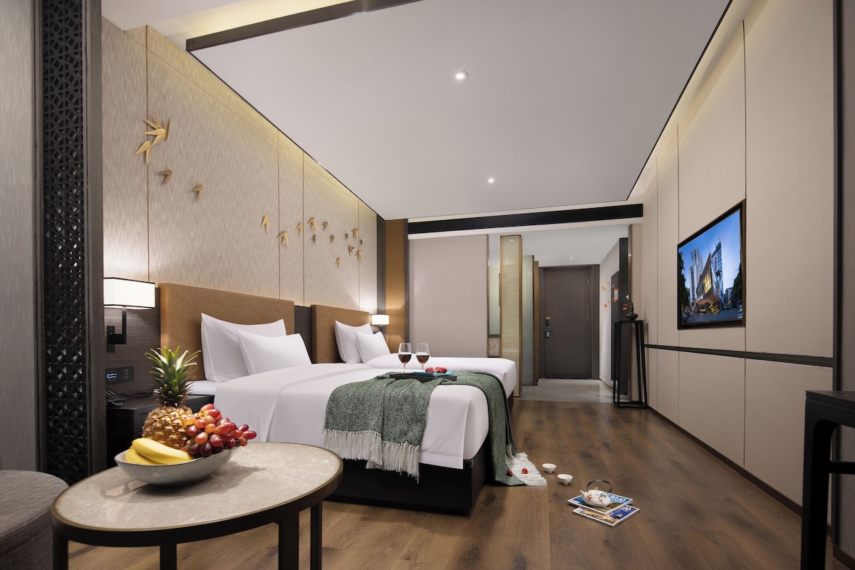 Una habitación de hotel moderna con una cama grande, una mesa de comedor con fruta, un televisor y vistas a la ciudad.