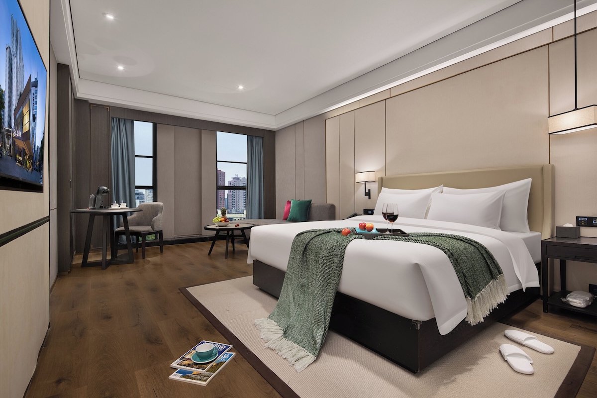 Habitación de hotel moderna con una cama grande, un escritorio, una silla, una mesita con fruta y vistas a la ciudad.