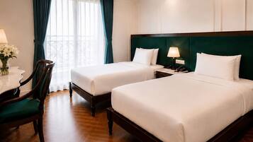 Ein Hotelzimmer mit zwei Einzelbetten, einem Schreibtisch mit Lampe, einem Sessel und Blick ins Freie durch ein Fenster mit Vorhängen.
