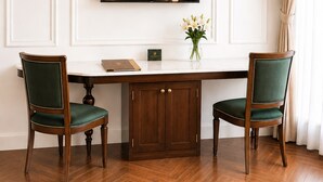 A room with a wooden herringbone floor, a white wall with a flat-screen TV mounted above a wooden table, two green upholstered chairs, and a vase with white flowers on the table.