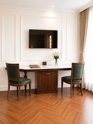 A room with a wooden herringbone floor, a white wall with a flat-screen TV mounted above a wooden table, two green upholstered chairs, and a vase with white flowers on the table.