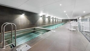 En moderne indendørs swimmingpool med aflangt design, omgivet af flisegulv og vægge, samt metalgelændere.
