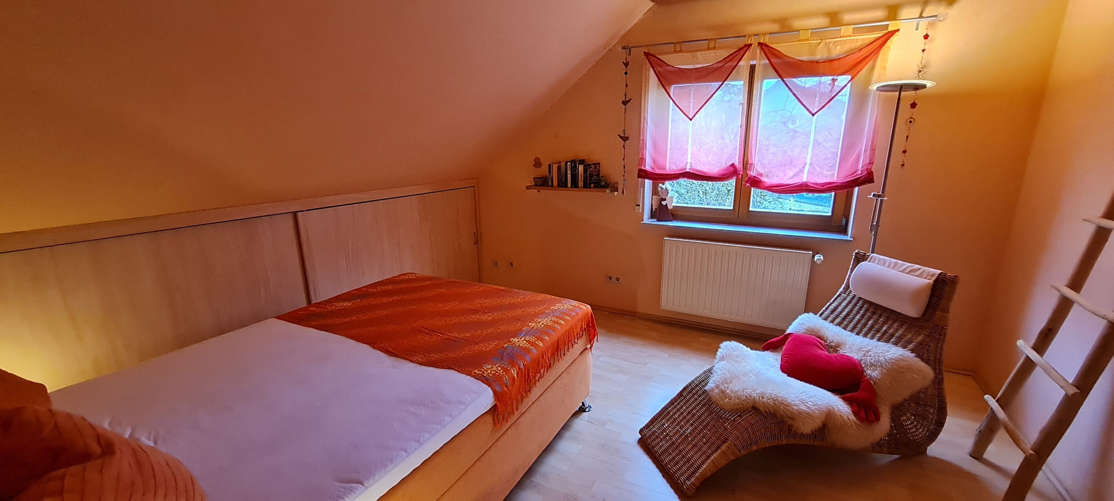 A bedroom with two beds, a window with curtains, a radiator, and a small shelf with books.