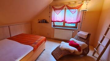 A bedroom with two beds, a window with curtains, a radiator, and a small shelf with books.