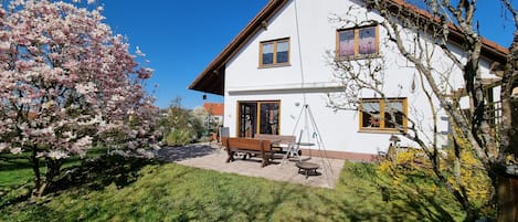 A two-story house with a patio, a swing set, and a garden with blooming trees.