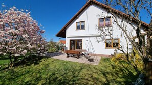 A two-story house with a patio, a swing set, and a garden with blooming trees.