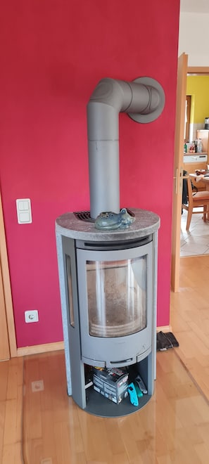 A compact wood stove with a grey chimney, a ceramic pot on top, and a power outlet on the wall.