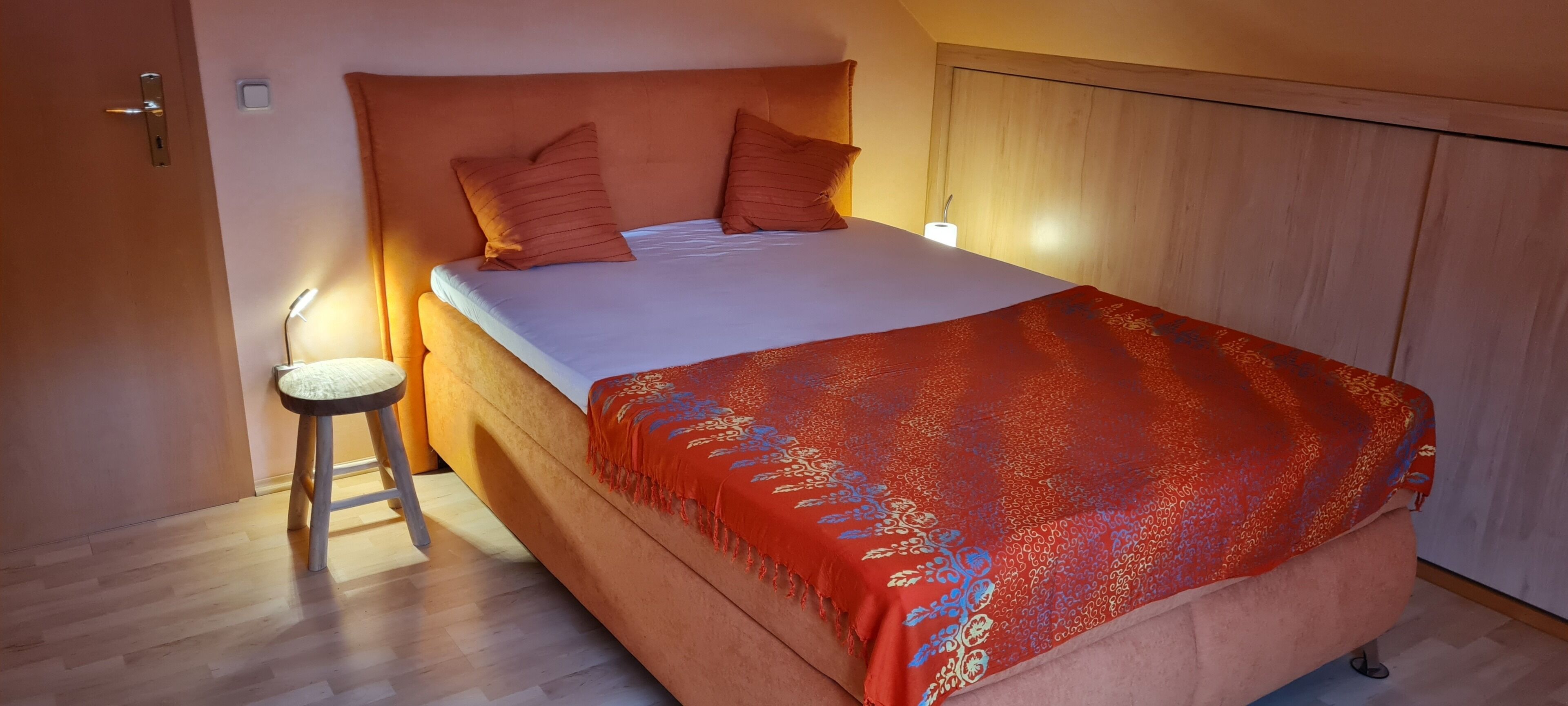 Een enkel bed met hoofdeinde, twee kussens, een nachtkastje met een lamp en een rood geborduurde deken.