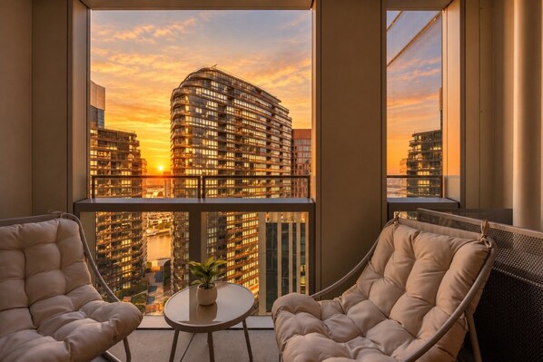 Ein Balkon mit zwei gepolsterten Stühlen, einem kleinen Tisch mit einer Pflanze und Blick auf hohe Gebäude bei Sonnenuntergang.