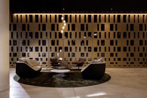 Eine Lobby mit modernem Design, ausgestattet mit einer runden Sitzecke, zwei Sofas, einem Couchtisch, einer dekorativen Leuchte und einer Wand mit zahlreichen kleinen Fenstern.