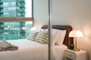A hotel room with a bed, bedside lamp, and a view of a cityscape through a window. - Stylish Modern 2B Apt w/ Pool, Gym, View & Library (Docklands)
