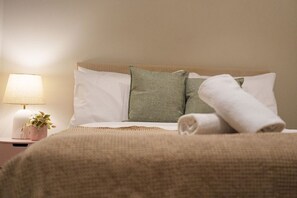Ein ordentlich bezogenes Bett mit einem aufgerollten Handtuch, einer Nachttischlampe und einem kleinen Topfpflanzen auf einem Nachttisch.