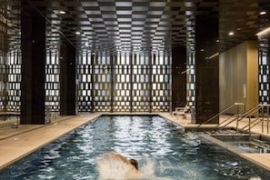 An indoor swimming pool with a checkered ceiling, clear glass walls, and a visible water surface. - Stylish Modern 2B Apt w/ Pool, Gym, View & Library (Docklands)