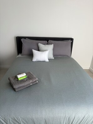 Ein ordentlich bezogenes Bett mit grauer Tagesdecke, Kissen und einem gefalteten Handtuch mit Pflegeprodukten.