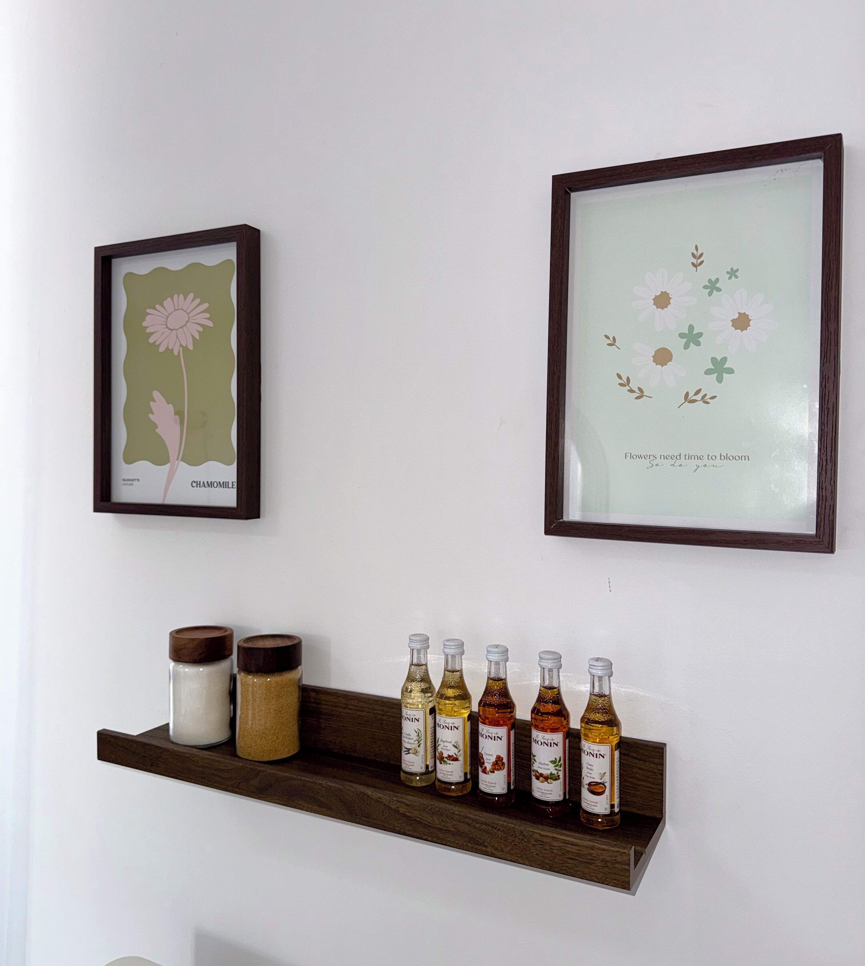 Un mur orné de deux reproductions botaniques encadrées et d’une étagère en bois où sont disposées des bouteilles et des bocaux.